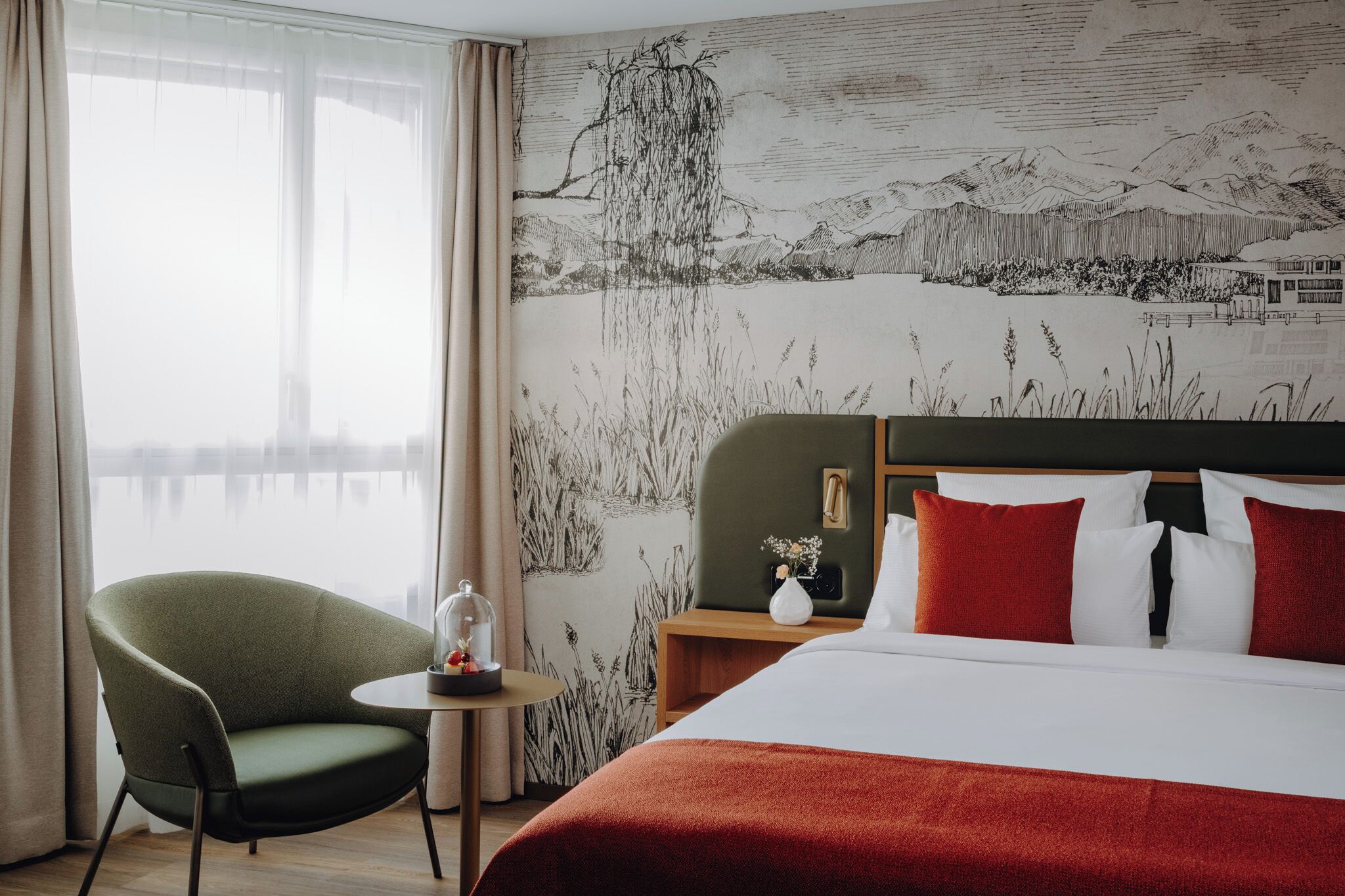 A hotel room with a bed, a green armchair, a small table with a vase, and a wall mural.