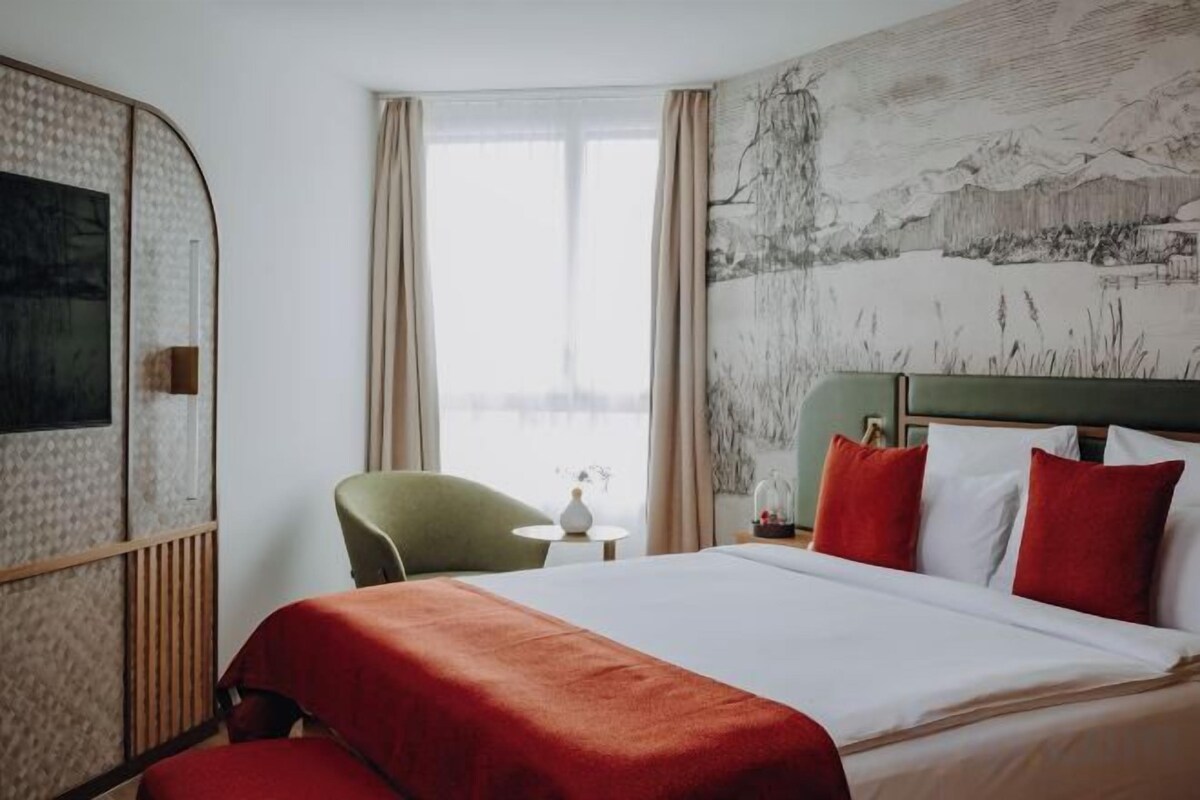 A hotel room with a bed, a green chair, a small table, and a painting on the wall.