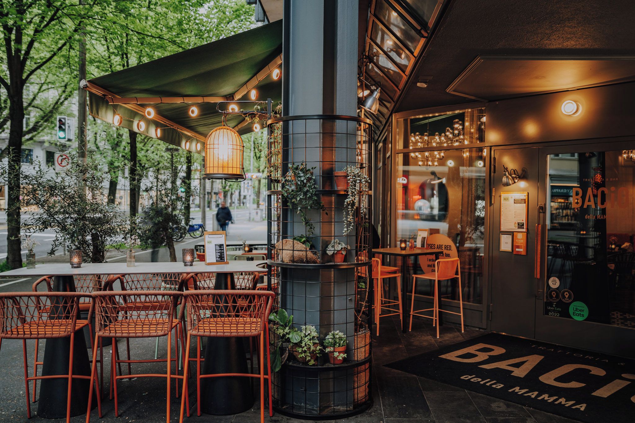 Un espace extérieur aménagé avec des chaises et des tables en osier, une colonne en métal ornée de plantes et une devanture de magasin portant l’enseigne « BACIO della Mamma ».
