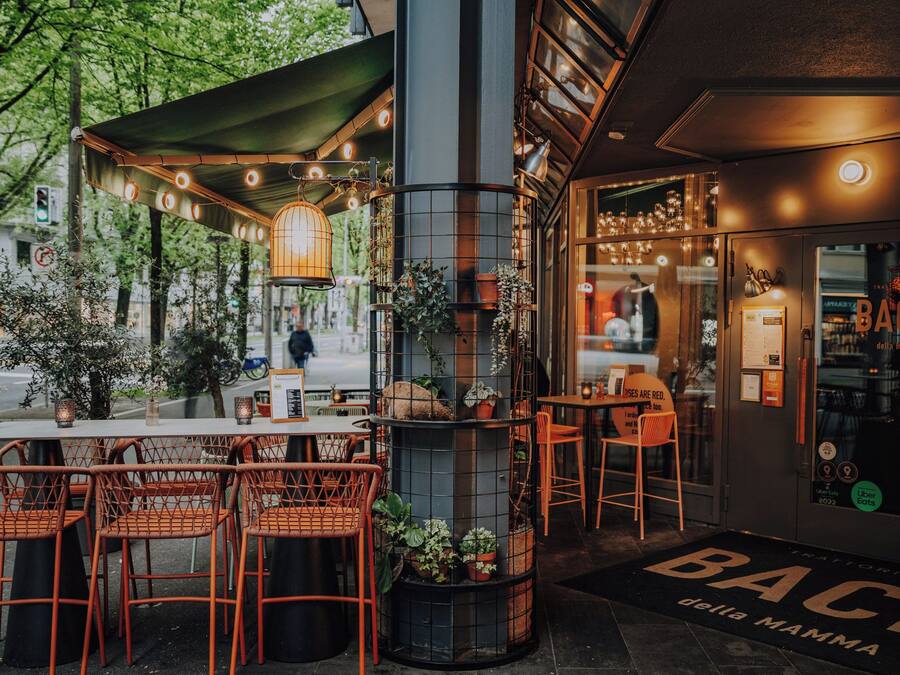 An outdoor seating area with wicker chairs and tables, a metal column with plants, and a storefront with a sign for 'BACIO della Mamma'.