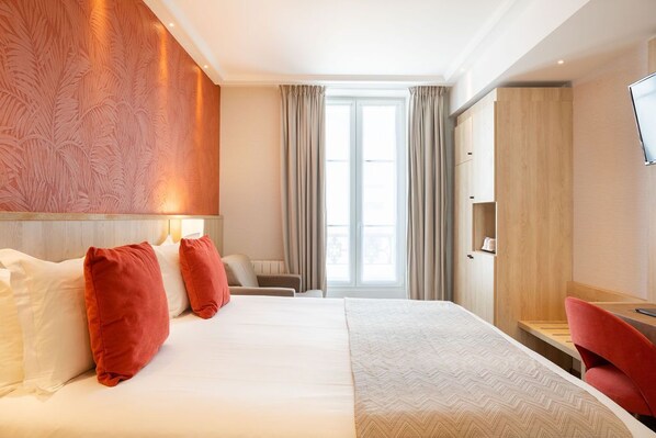 A hotel room with a large bed, a desk, a chair, a TV, and a wardrobe.
