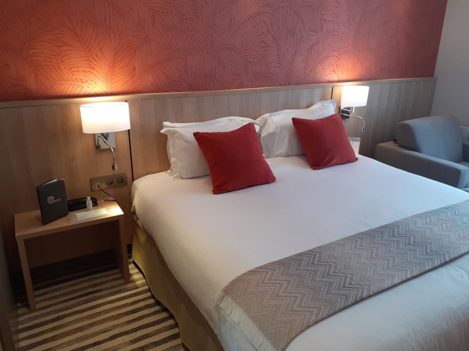 A hotel room with a large bed, two bedside lamps, a nightstand with a phone and a folder, and a chair.