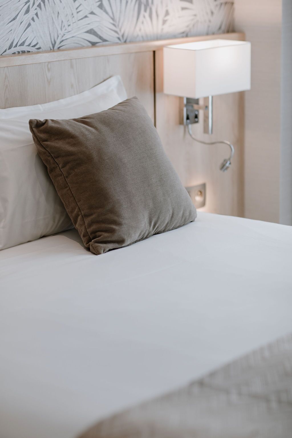 A neatly made bed with a single brown pillow and a white pillow, a wall-mounted lamp, and a wooden headboard with a leaf-patterned design.
