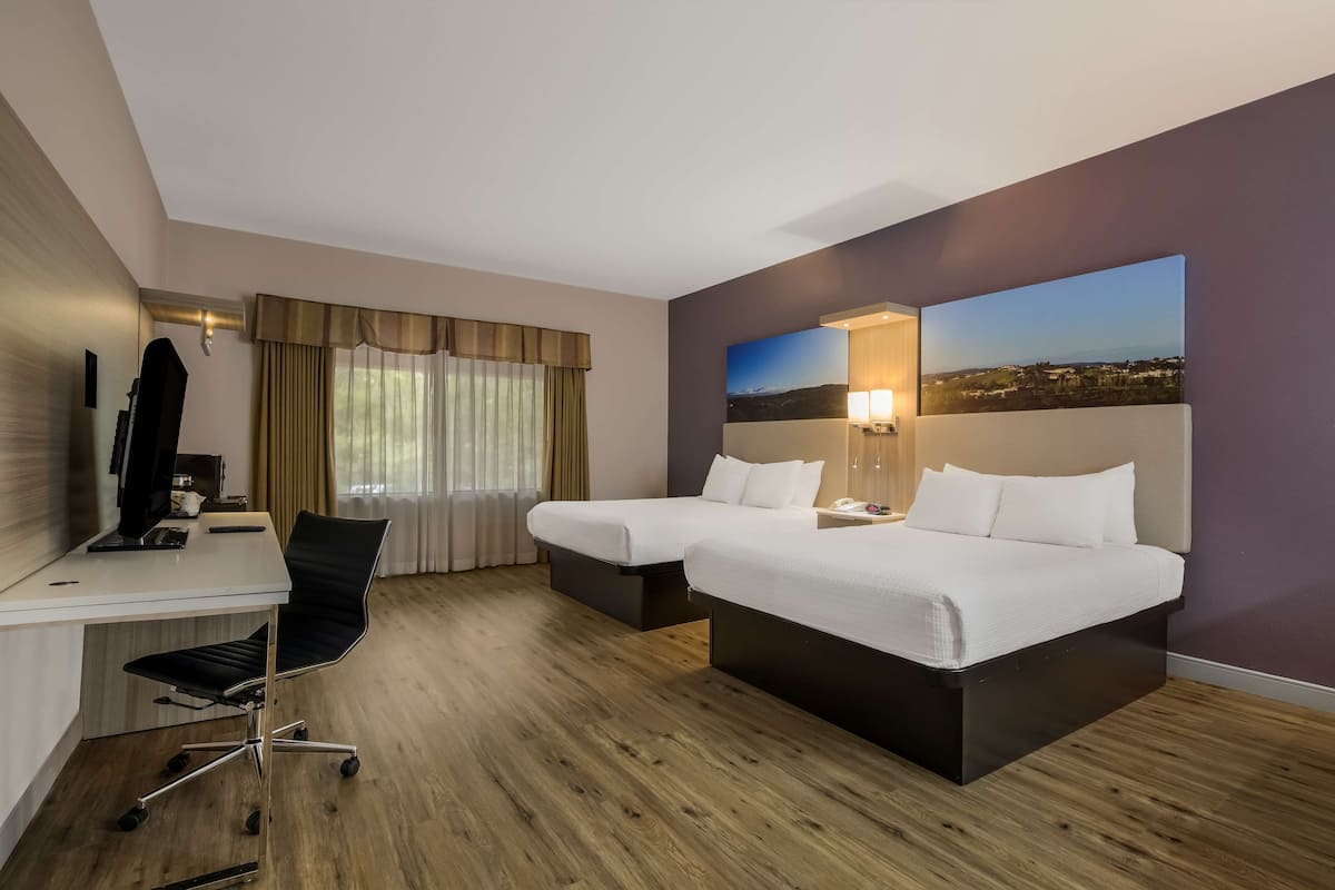 Habitación de hotel con dos camas, un escritorio con computadora, una silla y vista a un paisaje urbano.