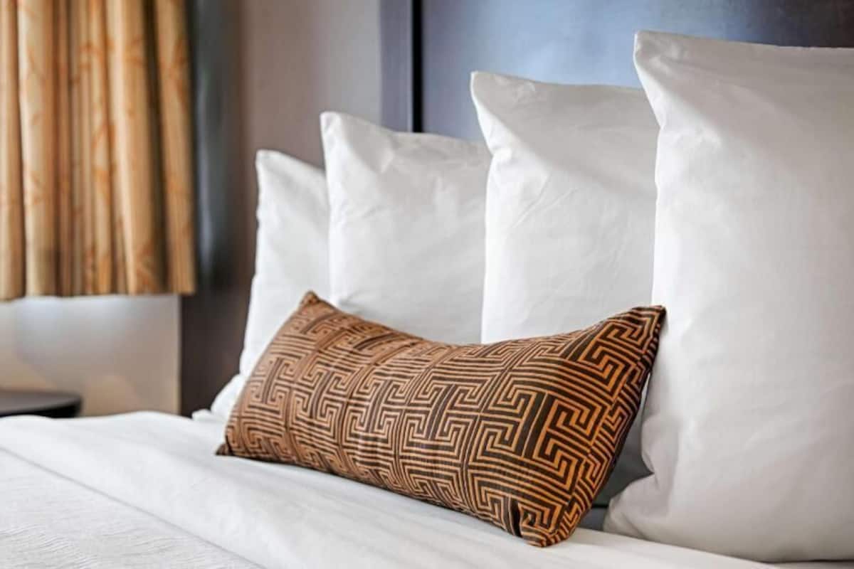 Una cama bien hecha con ropa blanca y una almohada estampada, un cabecero oscuro y una ventana con cortinas.