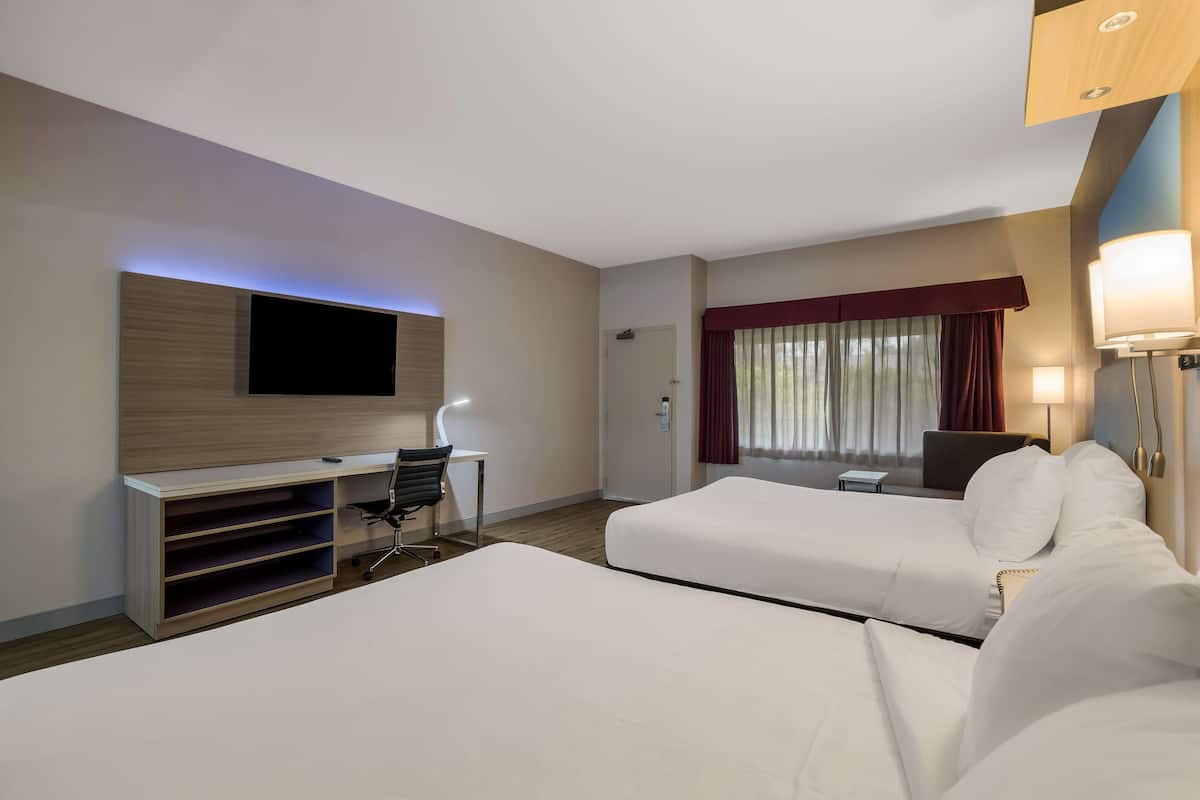 Habitación de hotel con dos camas, un escritorio con silla, televisor de pantalla plana montado en la pared, mesita de noche con lámpara y ventana con cortinas.