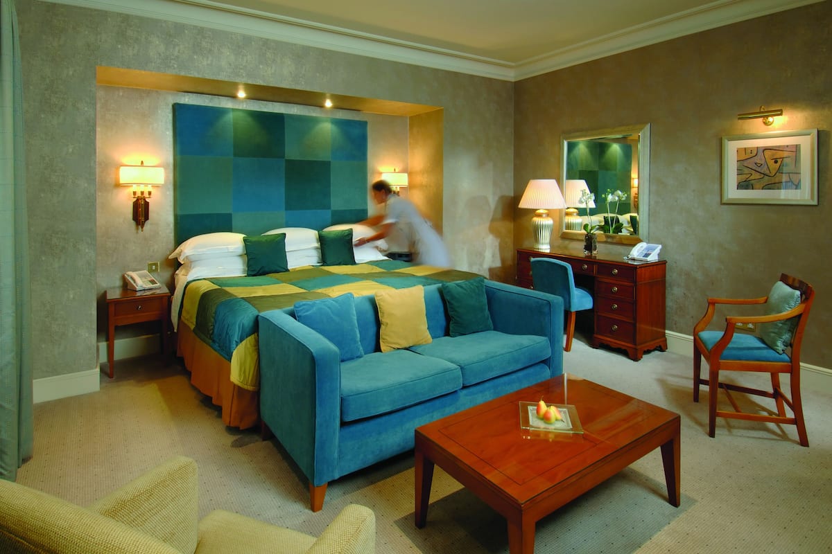 A hotel room with a large bed, a sofa, a wooden coffee table, a desk with a lamp, and a chair.