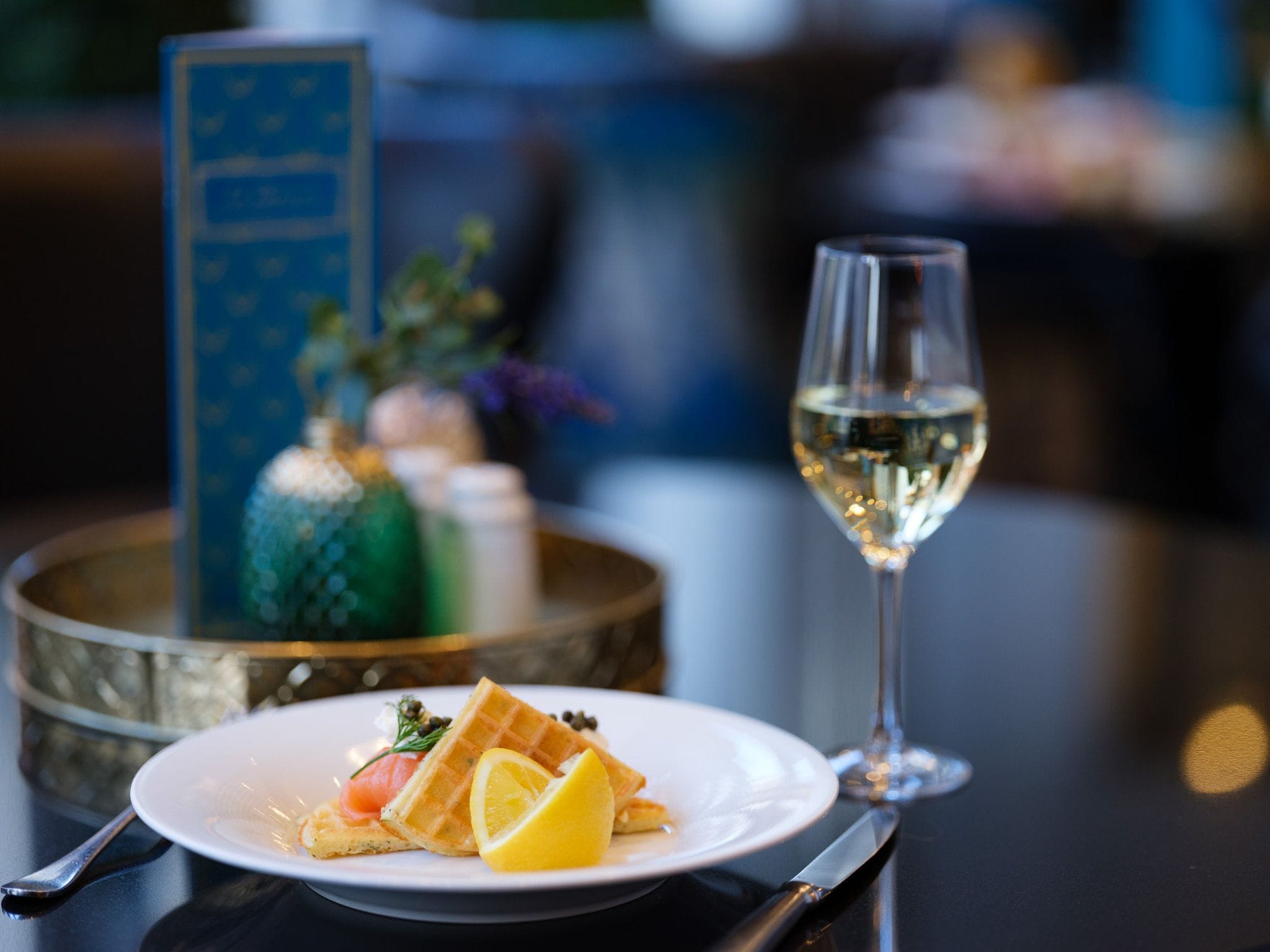 A plate with a waffle, lemon slice, and salmon, and a glass of white wine on a table.