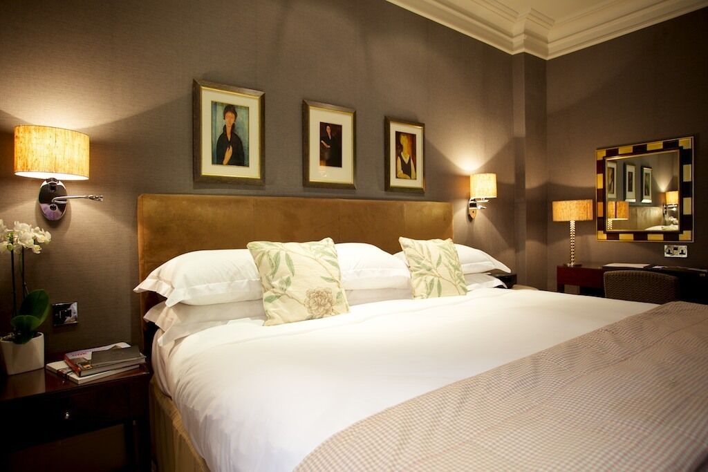 A hotel room with a large bed, two wall-mounted lamps, a bedside table with a lamp, a mirror, and framed artwork on the wall.