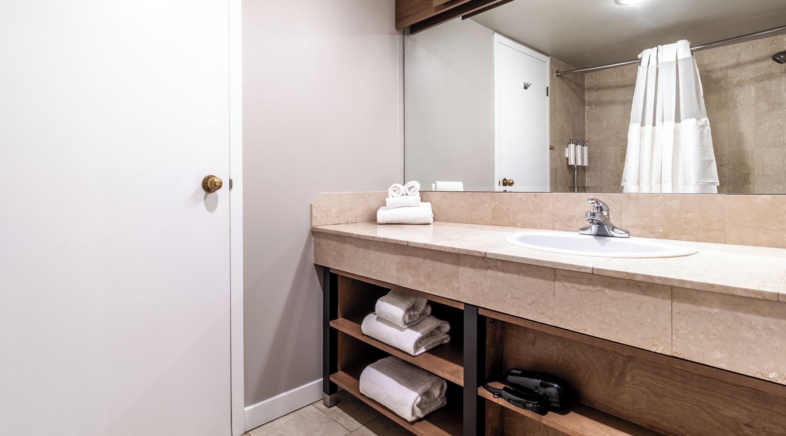 A modern bathroom with a beige marble countertop, a white sink, and a mirror above it. There are two shelves below the countertop holding folded towels.