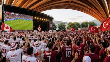 Un estadio lleno de aficionados, algunos con camisetas blancas y otros con camisetas rojas, con una pantalla gigante mostrando un partido de fútbol.