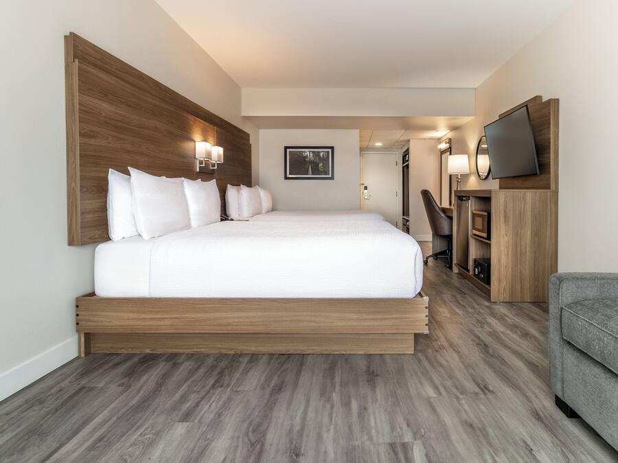 A modern hotel room with a large bed, wooden headboard, a flat-screen TV, a desk, and a grey sofa.