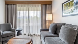 Una habitación de hotel moderna con un sofá gris, un reposapiés marrón, una mesita auxiliar y un ventanal con cortinas translúcidas.
