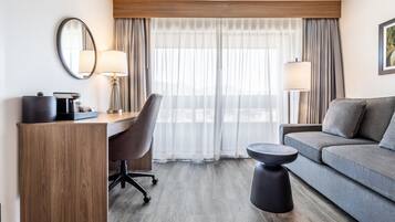 A hotel room with a wooden desk, a grey sofa, a round black ottoman, a chair, and a large window with sheer curtains.