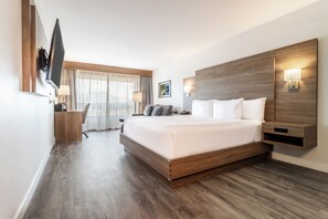 A modern hotel room with a large bed, wooden headboard, a flat-screen TV, a desk with a chair, and a window with curtains.