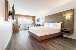 A modern hotel room with a large bed, wooden headboard, a flat-screen TV, a desk with a chair, and a window with curtains.