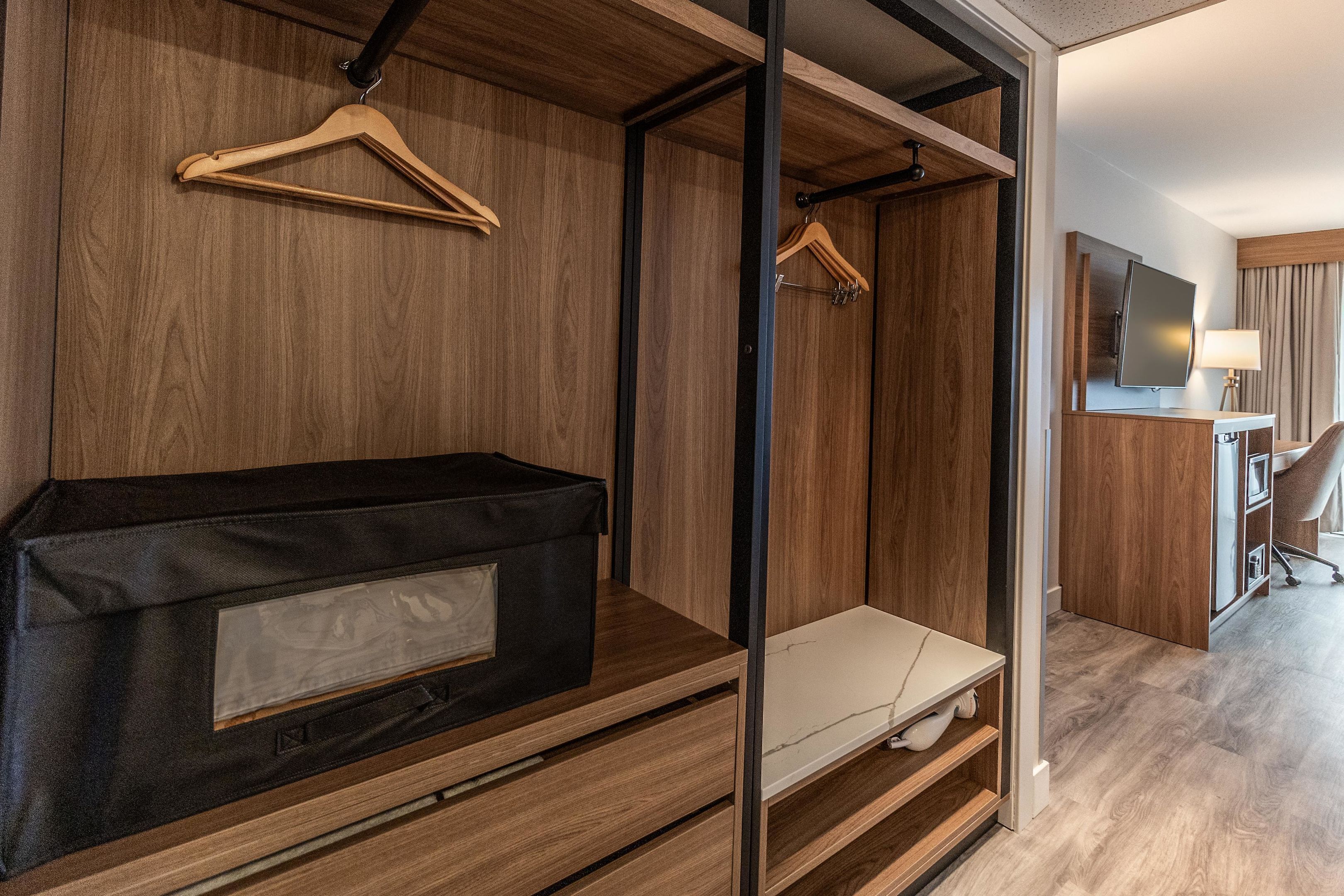 A hotel room with a wooden wardrobe, a bench, a desk with a lamp, and a television.