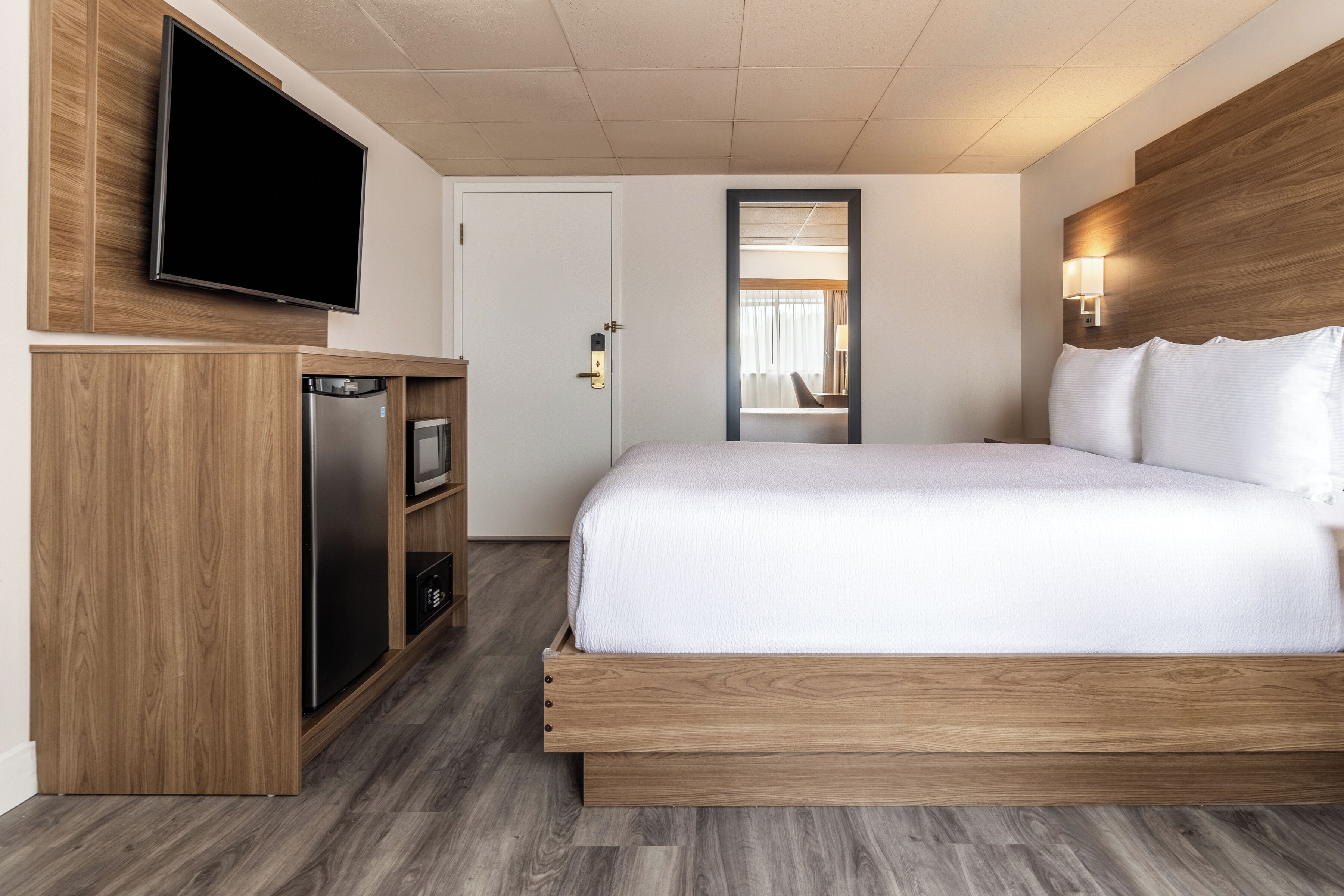 A hotel room with a bed, a TV mounted on the wall, a small kitchenette, and a mirror above the door.