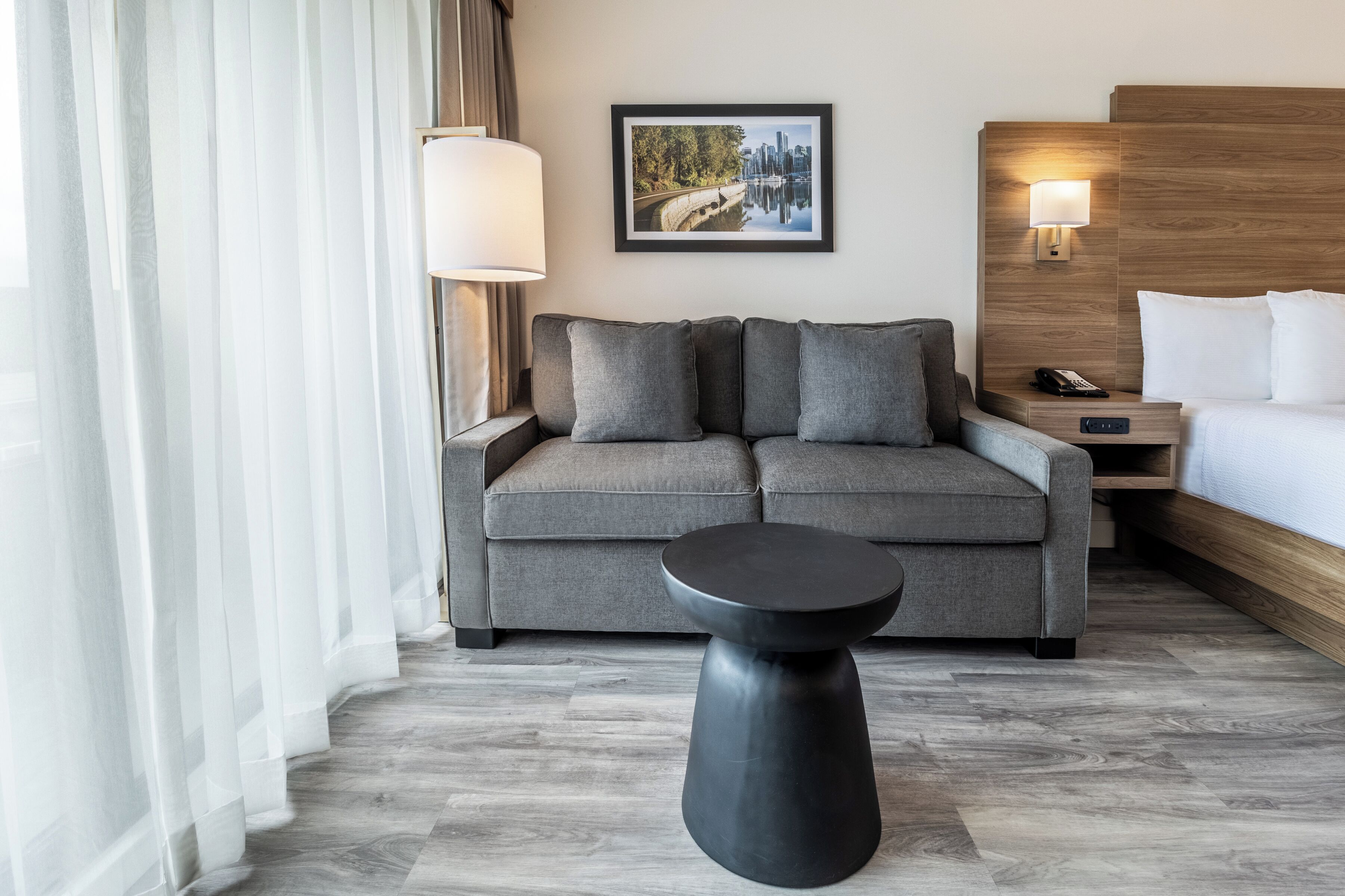 A modern hotel room with a grey sofa, a small black side table, a bed with white linens, a framed cityscape picture on the wall, and a wooden headboard.