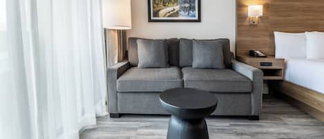 A modern hotel room with a grey sofa, a small black side table, a bed with white linens, a framed cityscape picture on the wall, and a wooden headboard.