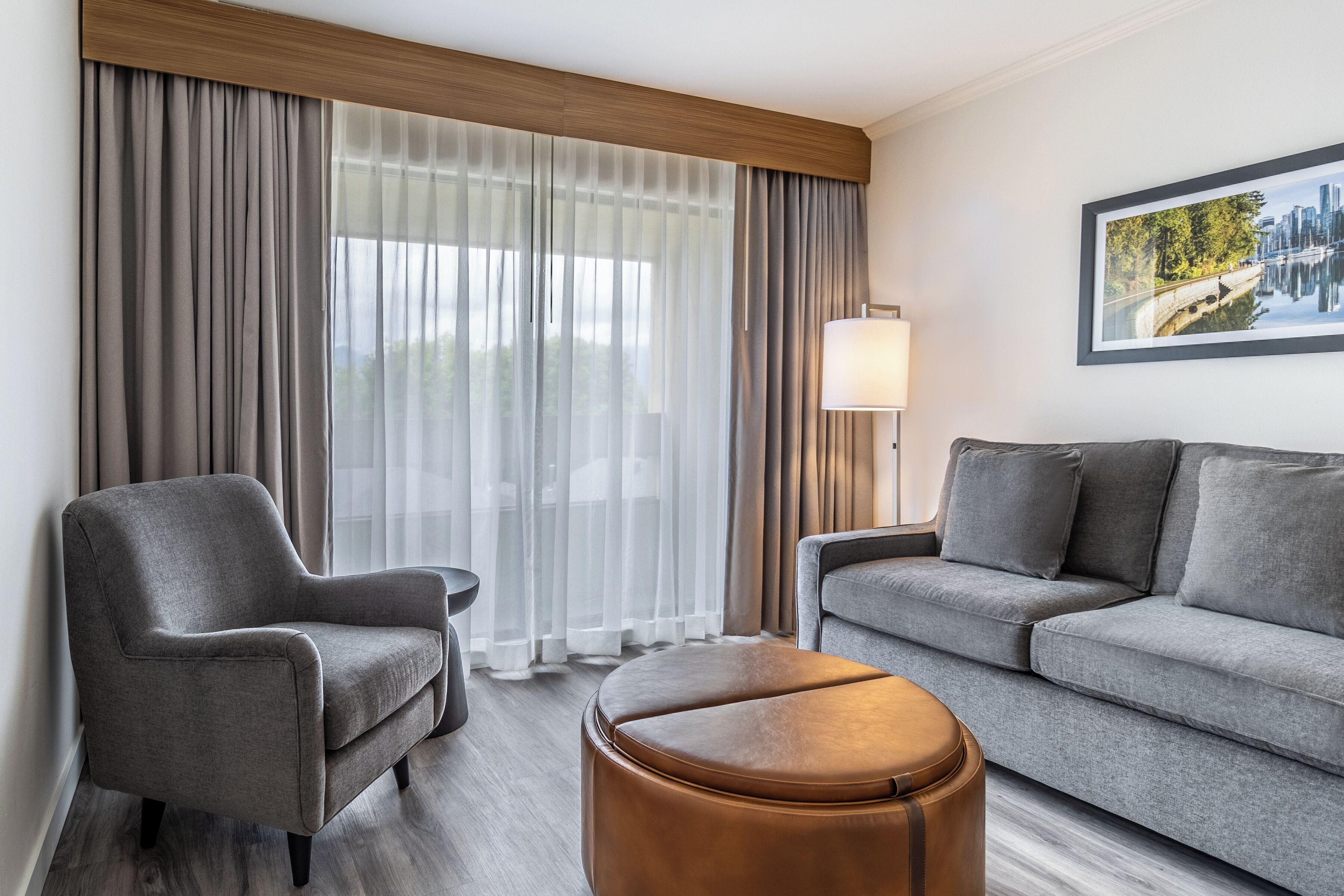 Una habitación de hotel moderna con un sofá gris, un sillón gris, un reposapiés de madera y un ventanal con cortinas translúcidas.