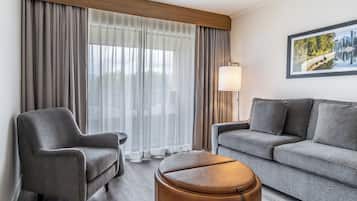 Una habitación de hotel moderna con un sofá gris, un sillón gris, un reposapiés de madera y un ventanal con cortinas translúcidas.