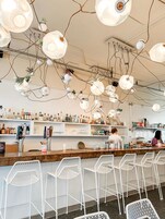Un bar con sillas de metal blanco, estantes con botellas y lámparas colgantes.