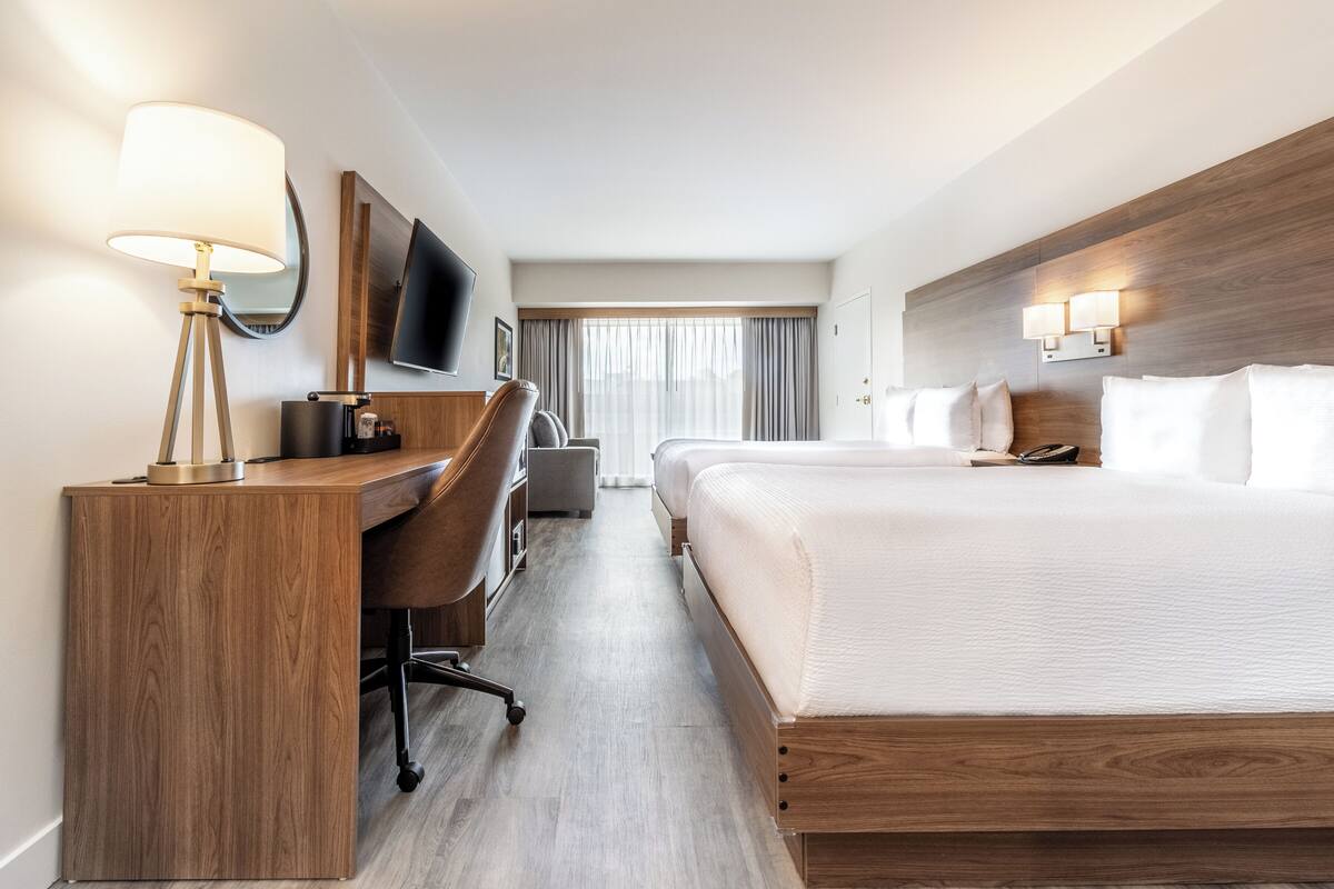 A hotel room with a wooden desk, a chair, a bed with white bedding, a TV mounted on the wall, and a window with curtains.