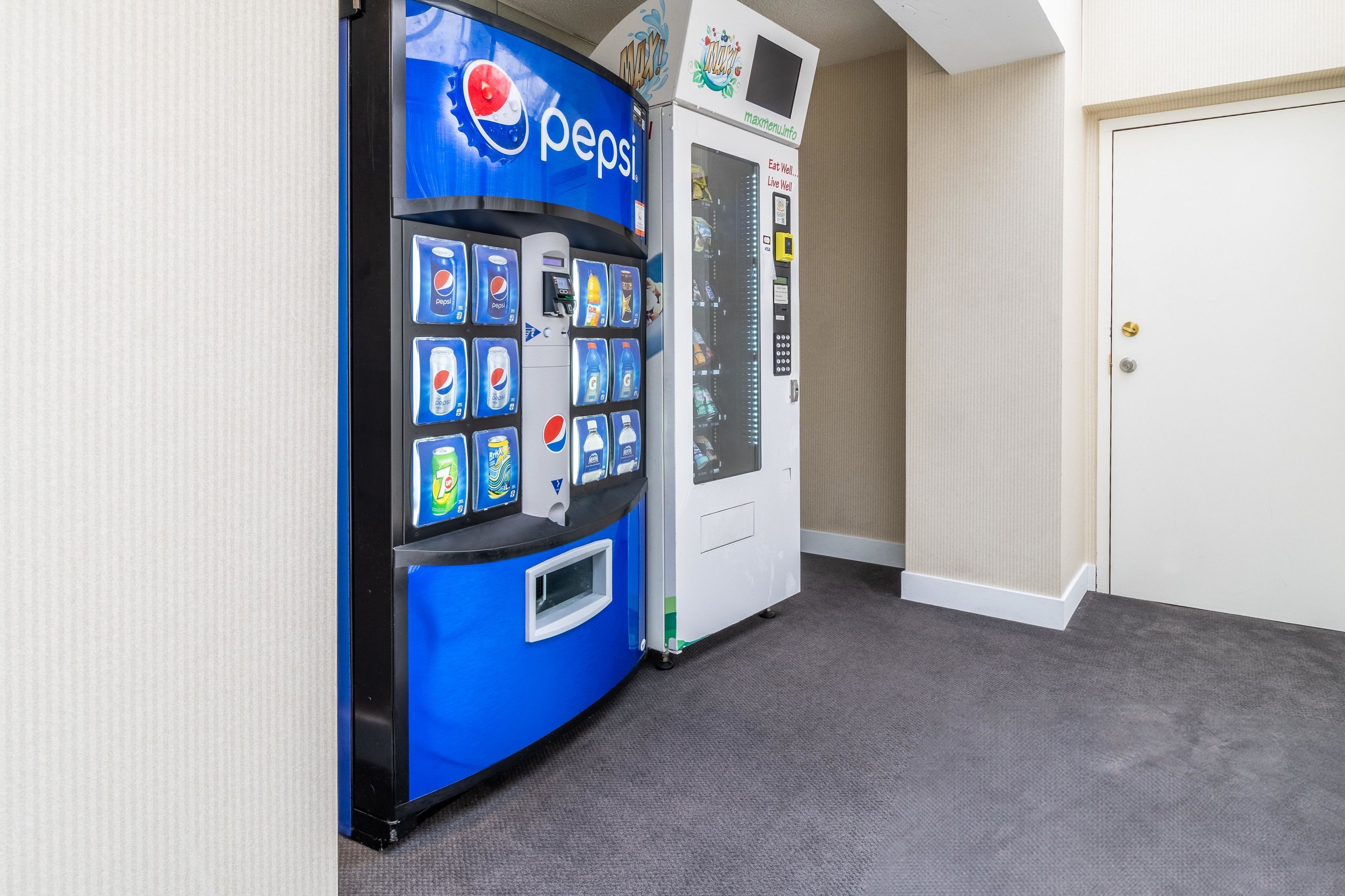 A vending machine with a Pepsi logo, a white machine with a digital display, and a closed door in a hallway.