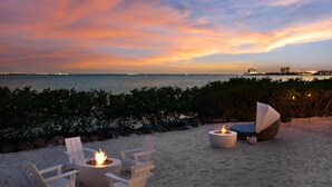 Un espace aménagé avec des chaises et un foyer, situé au bord de la plage, d’où l’on peut admirer l’océan et le panorama de la ville au coucher du soleil.