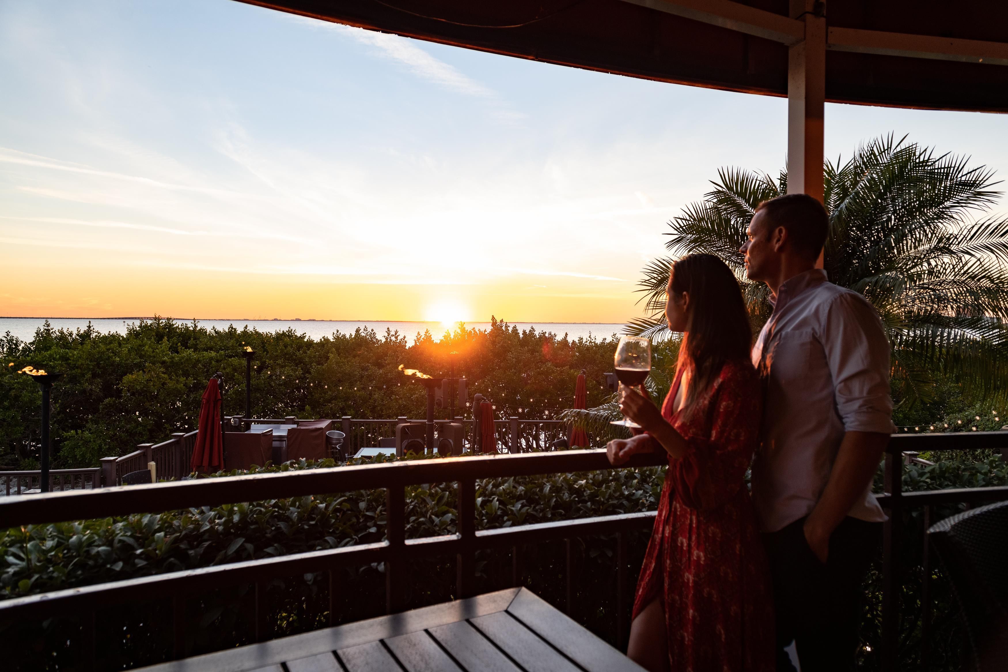 A couple standing on a balcony overlooking a sunset view with a body of water and greenery.