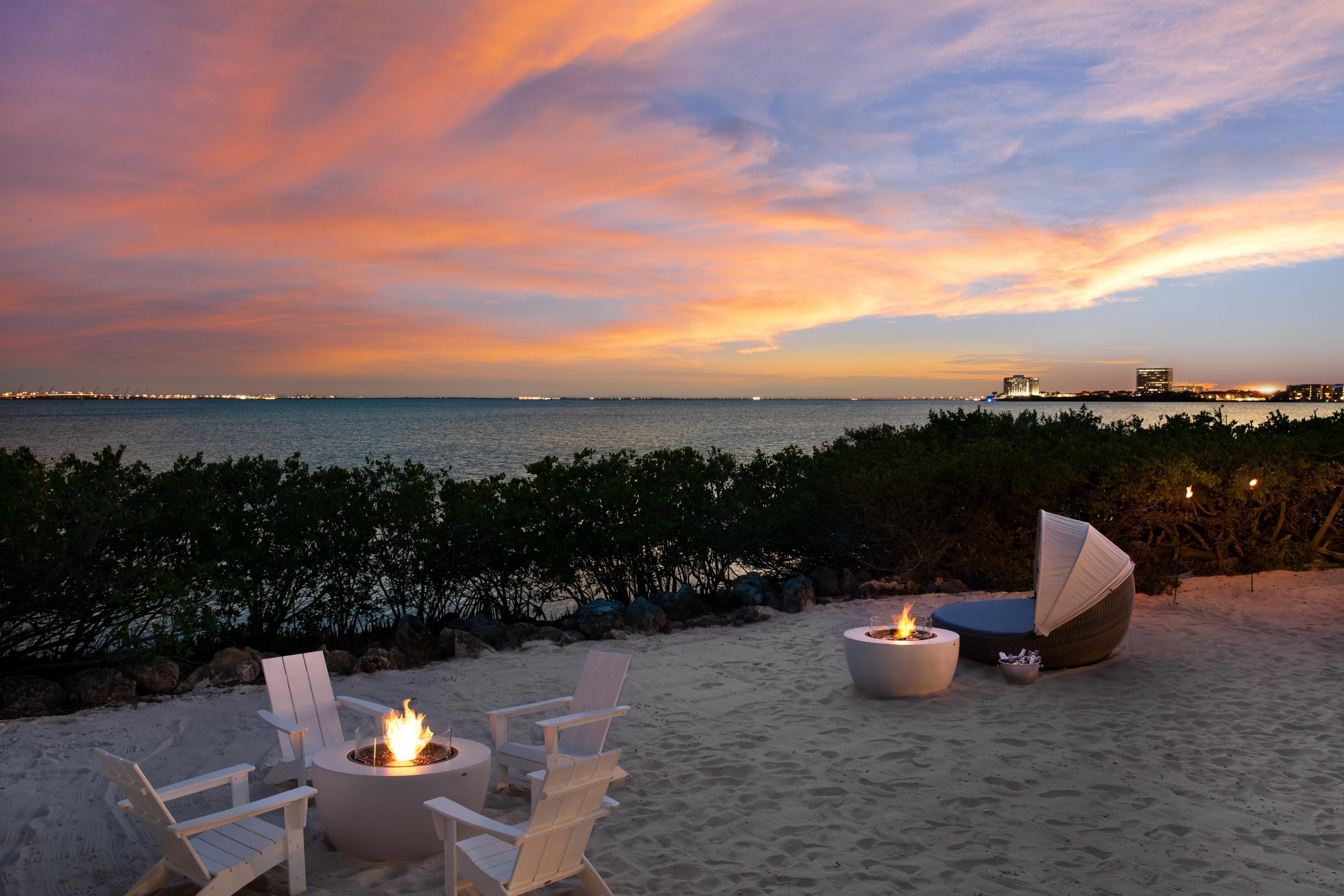 A beachside seating area with a fire pit, lounge chairs, and a view of the ocean and city skyline at sunset.