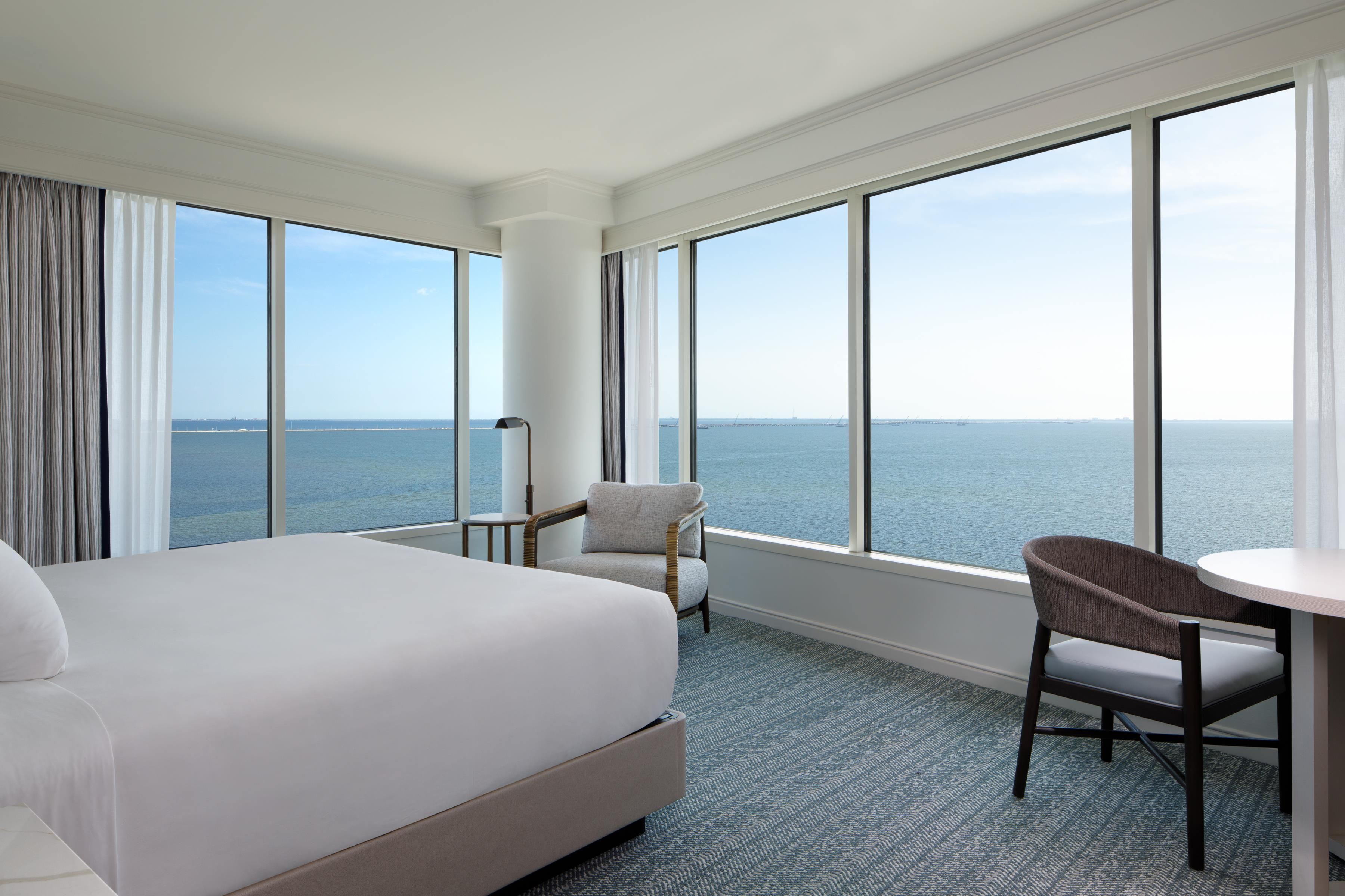 Une chambre d’hôtel avec un grand lit, une chaise, une petite table et une vue sur la mer grâce à de grandes fenêtres.