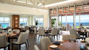 Un vaste espace dédié à la salle à manger, avec de nombreuses tables et chaises, de grandes fenêtres offrant une vue sur l’océan et un plafond haut avec des poutres apparentes.