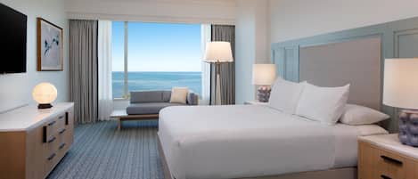A hotel room with a large bed, two bedside tables, a sofa, a TV, and a view of the sea.