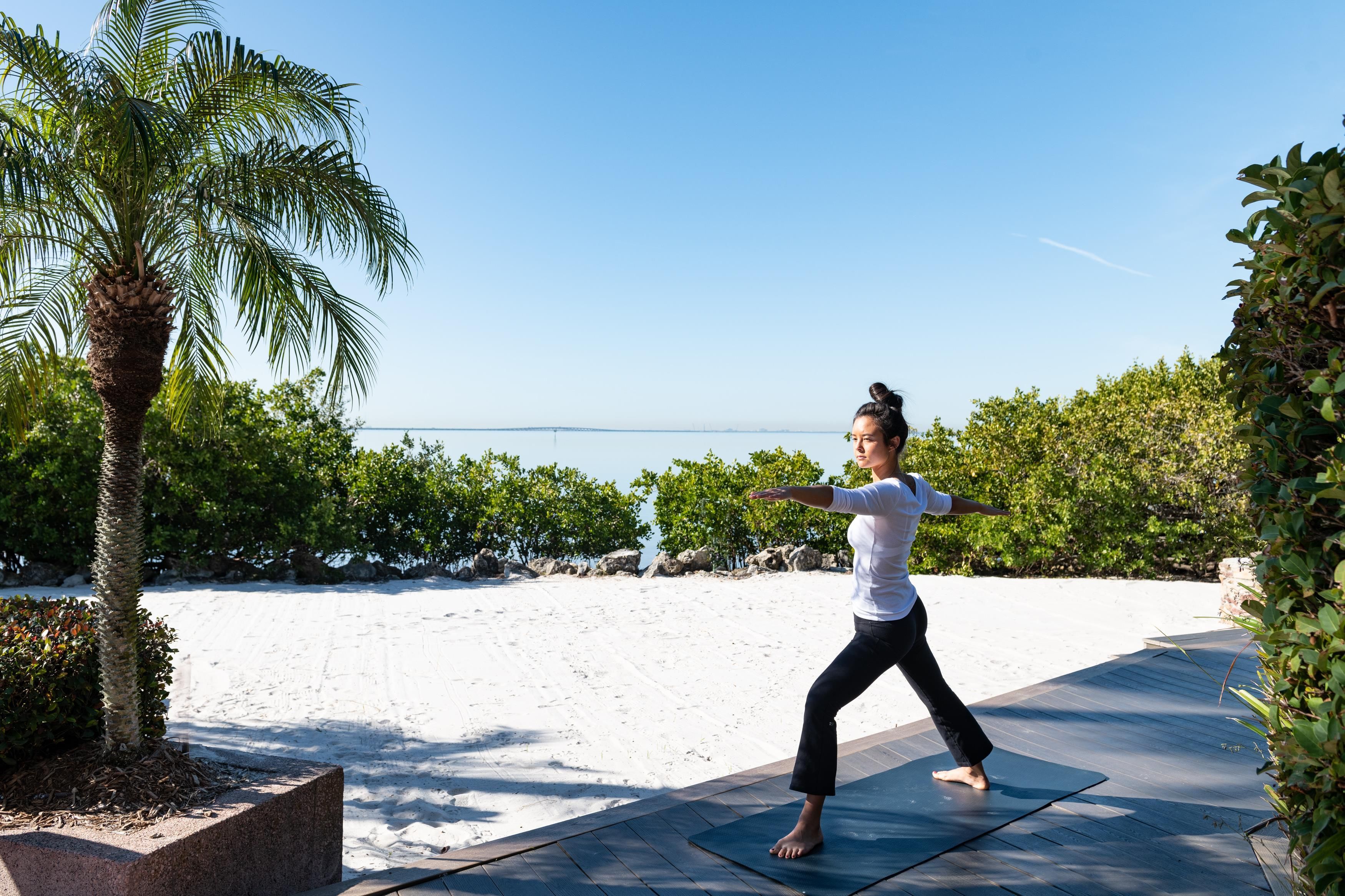 Iemand doet yoga op een mat, buiten, met een heldere lucht en een strand op de achtergrond.