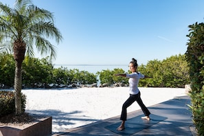 Una persona practicando yoga sobre una esterilla al aire libre, con un cielo despejado y una playa de fondo.