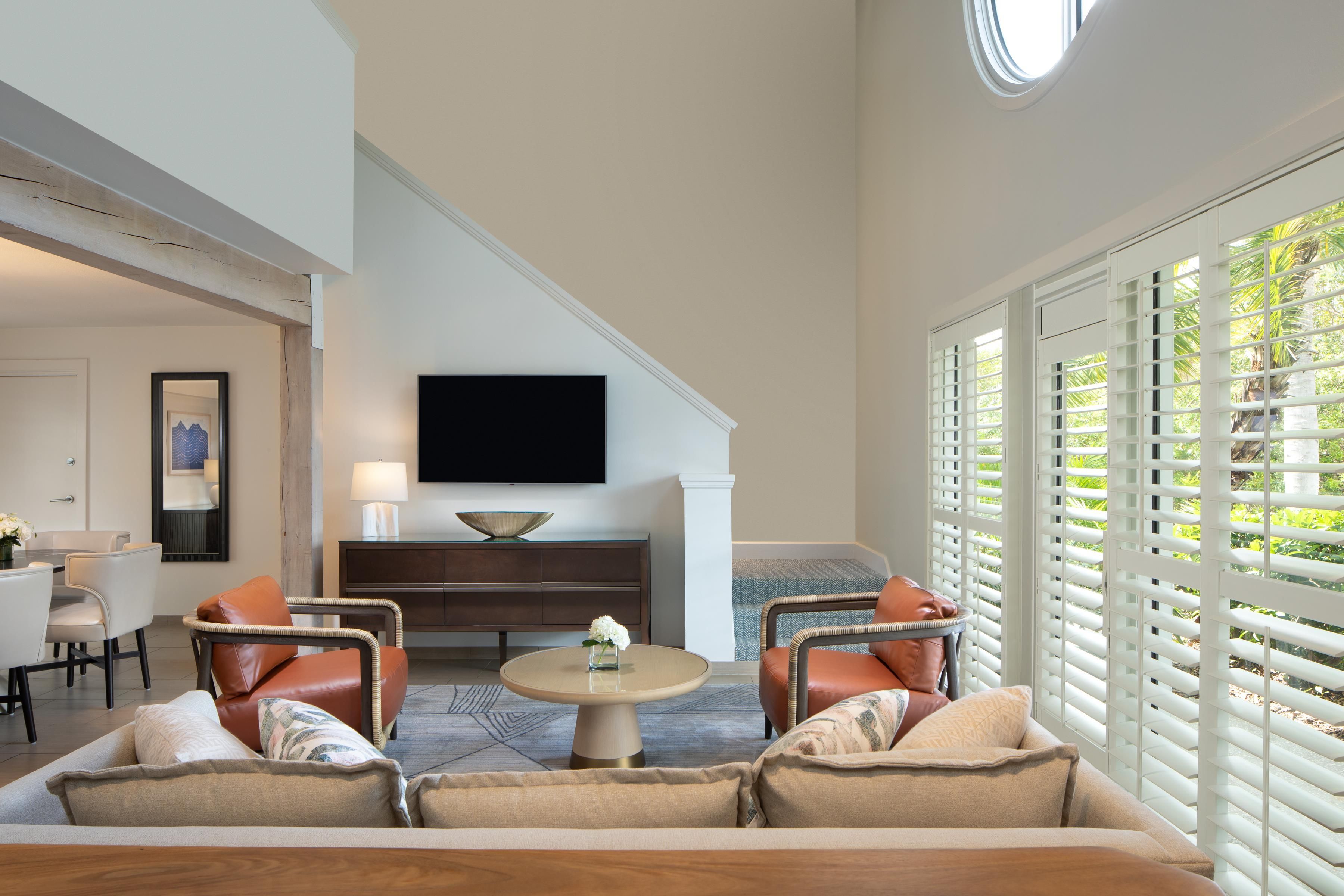 A modern living room with a sofa, chairs, a coffee table, a flat-screen TV, and a large window with blinds.