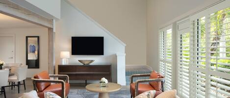 A modern living room with a sofa, chairs, a coffee table, a flat-screen TV, and a large window with blinds.