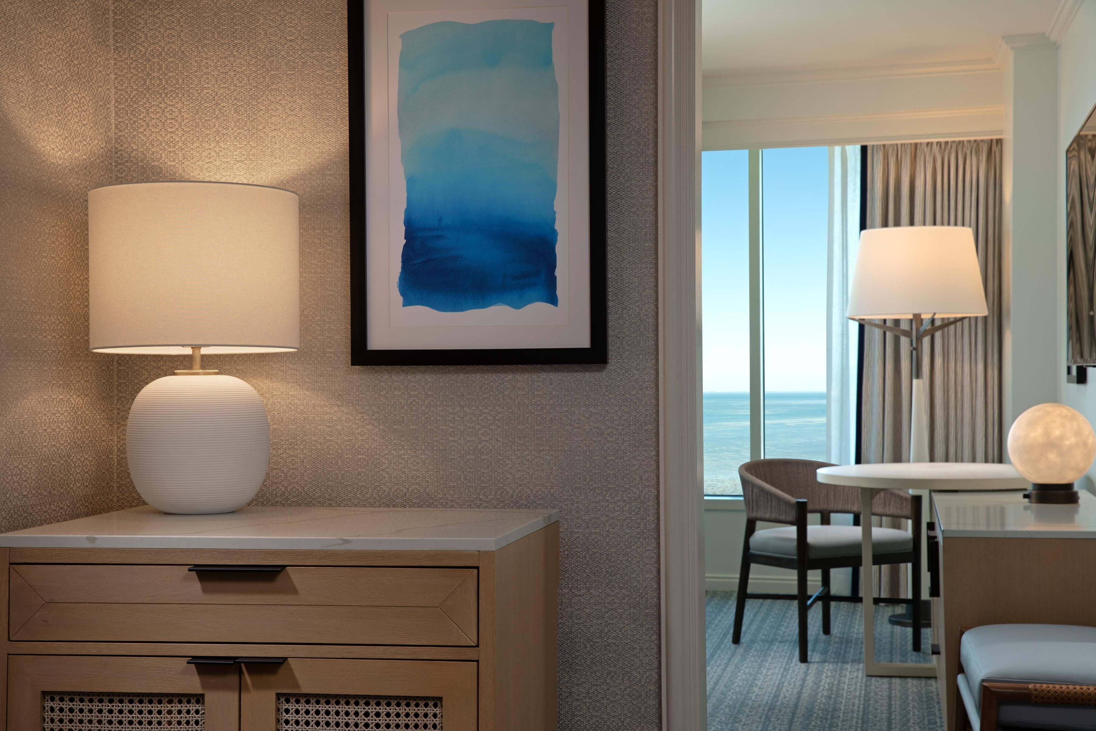 A hotel room with a bed, a desk, a chair, a lamp, a framed abstract painting, and a view of the sea through the window.