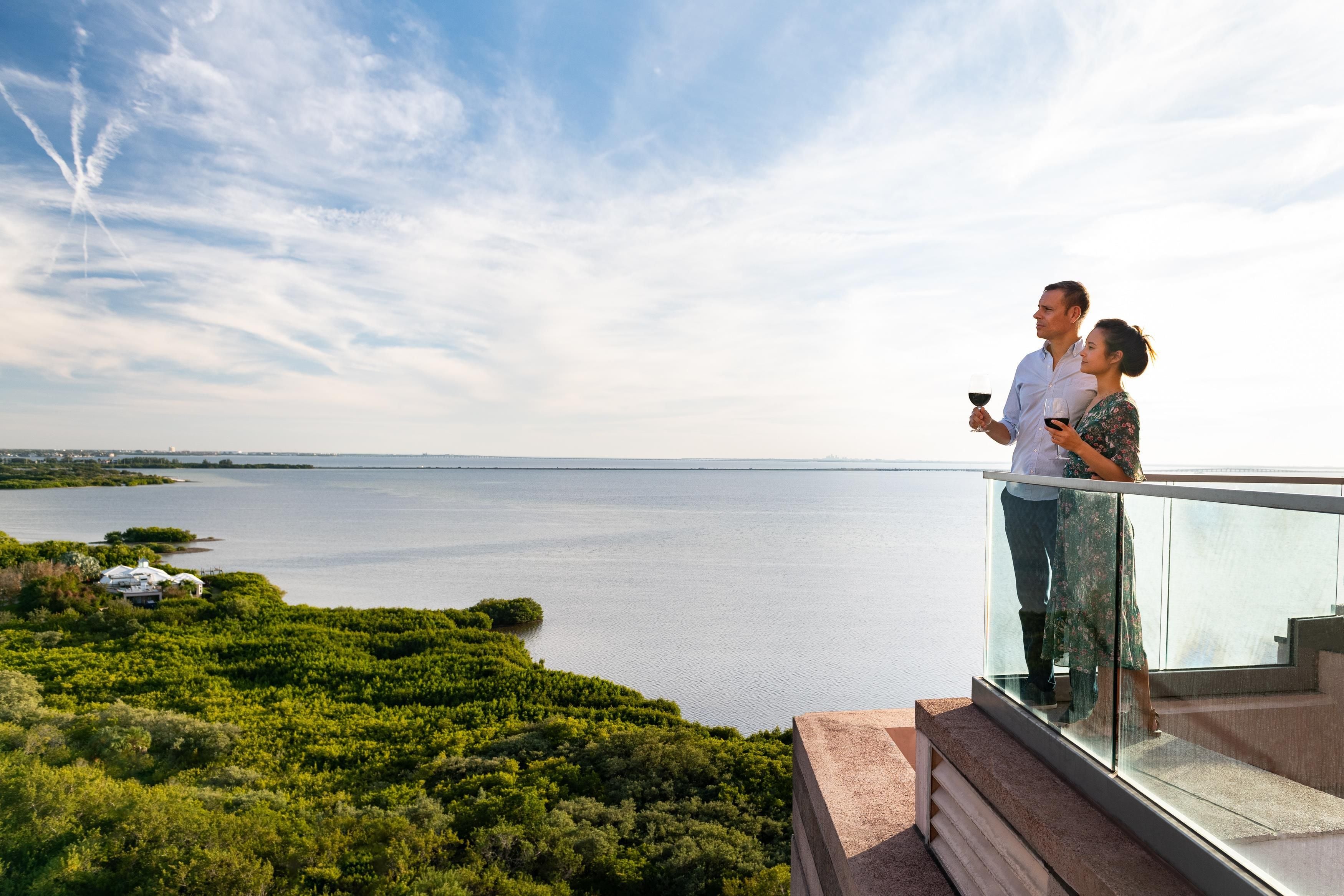 A couple standing on a balcony overlooking a body of water with a clear sky.