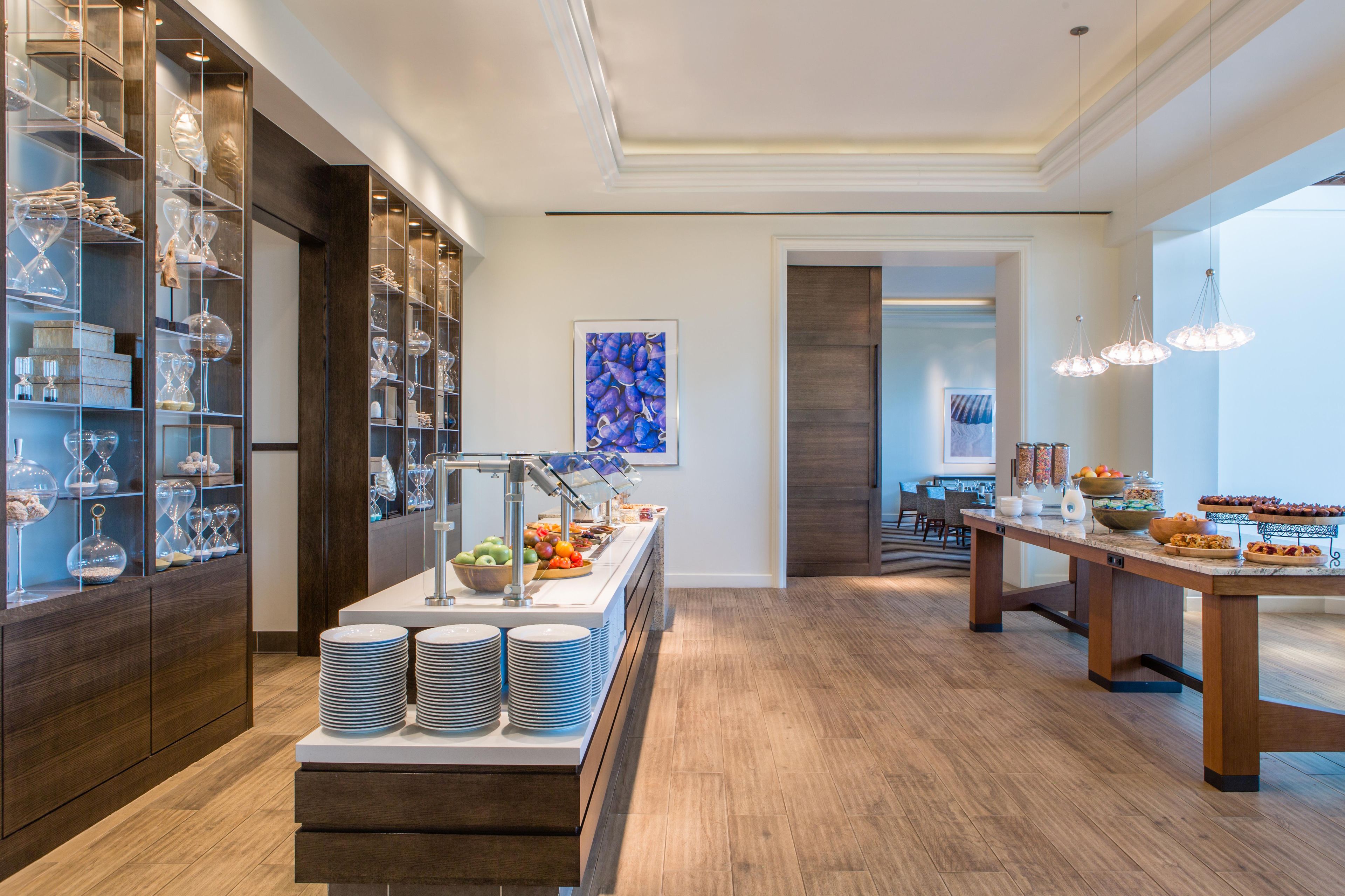 A modern hotel breakfast area with a buffet setup, a dining area with a long table, and a view into another room.