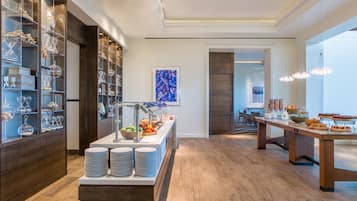 A modern hotel breakfast area with a buffet setup, a dining area with a long table, and a view into another room.
