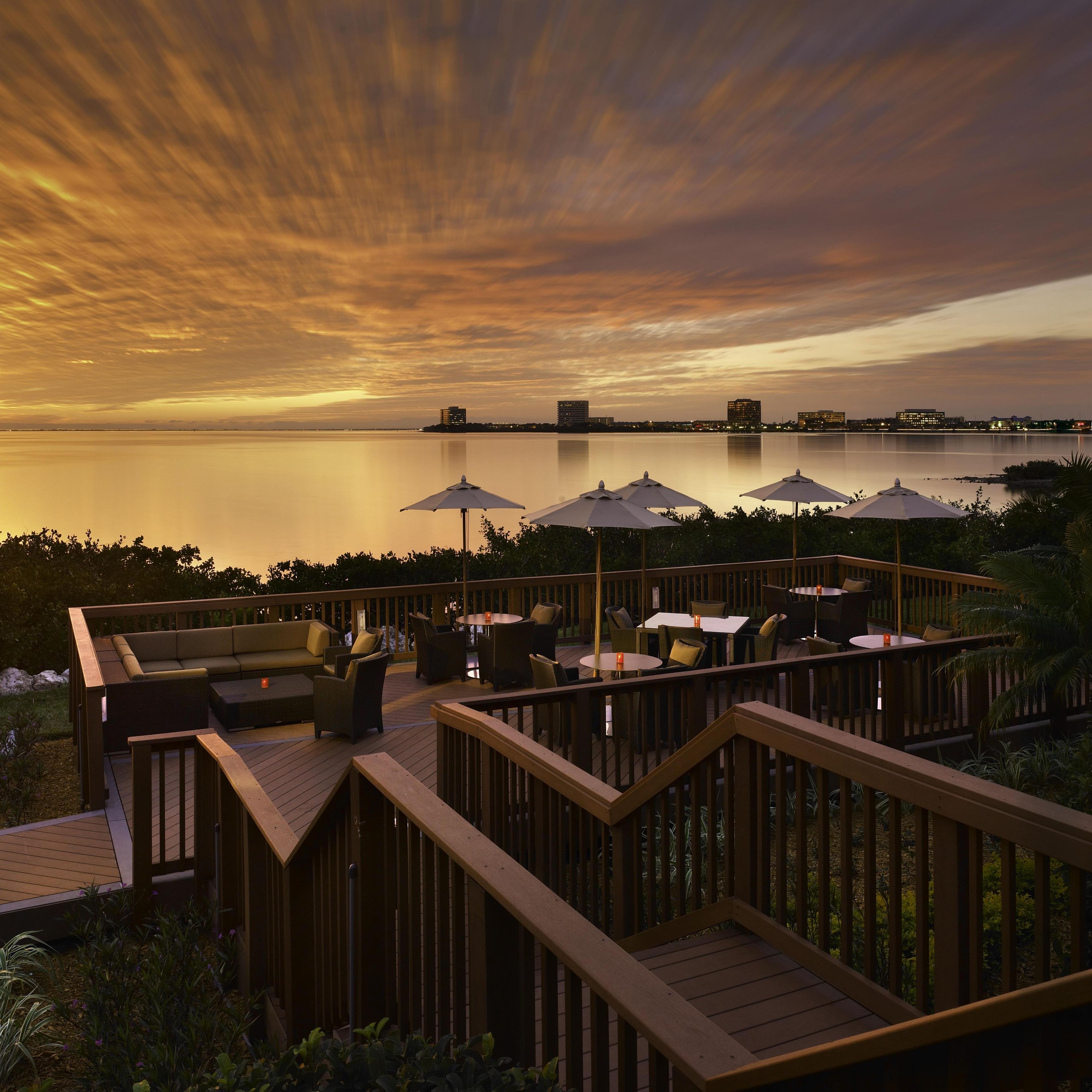 A deck with outdoor seating and umbrellas overlooking a calm body of water at sunset.