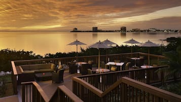 A deck with outdoor seating and umbrellas overlooking a calm body of water at sunset.
