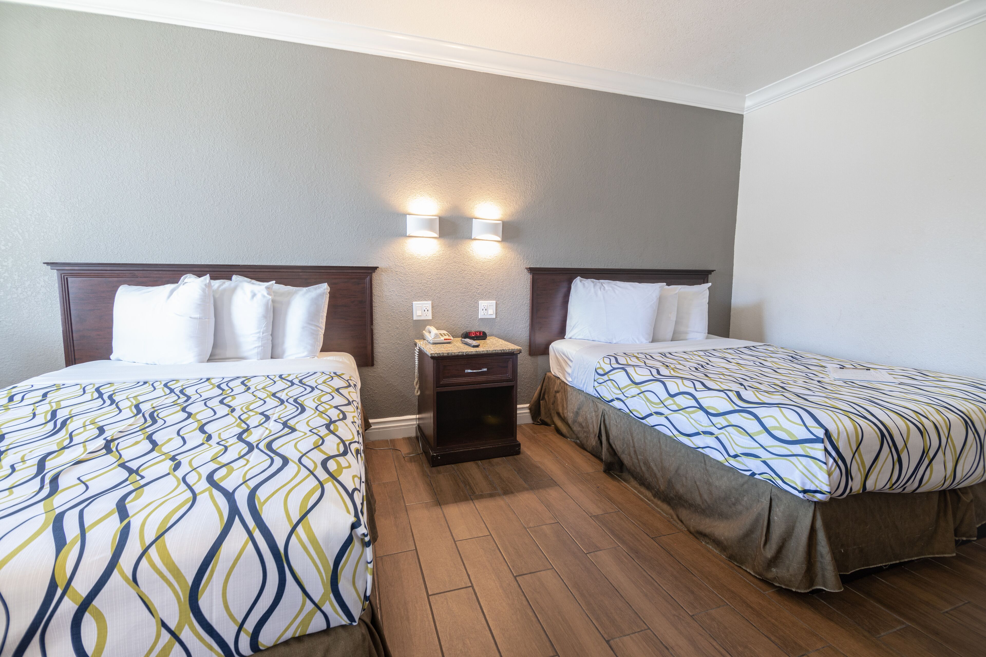 A hotel room with two beds, a nightstand with a phone and clock, and two wall-mounted lights.