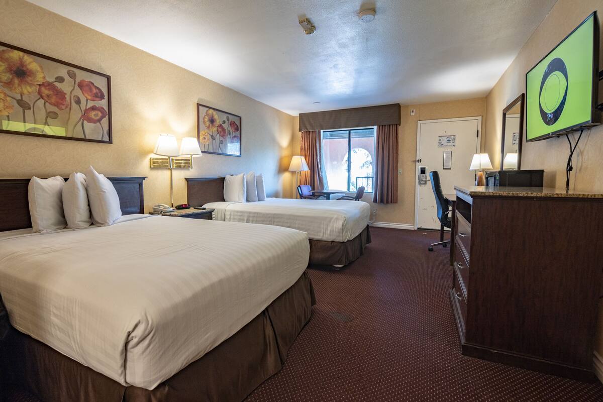 A hotel room with two beds, a desk, a chair, a lamp, and a TV.