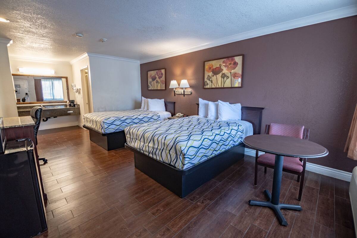 A hotel room with two beds, a small table, and a chair. There is a desk with a chair and a mirror.