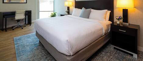 A hotel room with a large bed, two bedside tables with lamps, a desk with a chair, and a window.