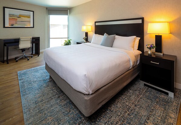A hotel room with a large bed, two bedside tables with lamps, a desk with a chair, and a window.